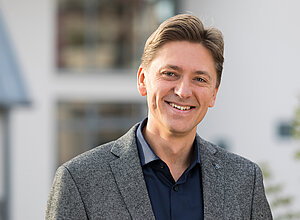 Partnermanager Jurij Deperschmidt steht lächelnd mit blauem Hemd und grauem Sakko vor einem Gebäude.