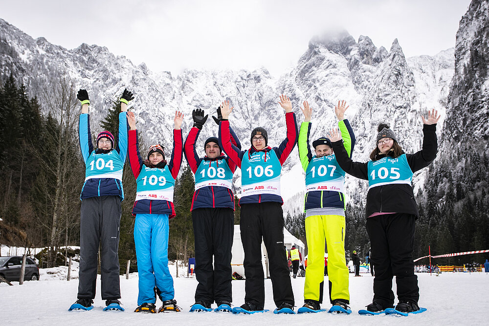 Sechs Personen stehen in einer verschneiten Berglandschaft. Sie tragen bunte Skihosen und Trikots mit Nummern und haben die Arme gehoben.