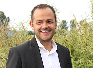 Partnermanager Johannes Eisele steht in Anzug lächelnd vor einem Busch.