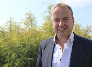 Partnermanager Dominik Baumann steht lächelnd in Anzug in der Sonne vor einem Busch.