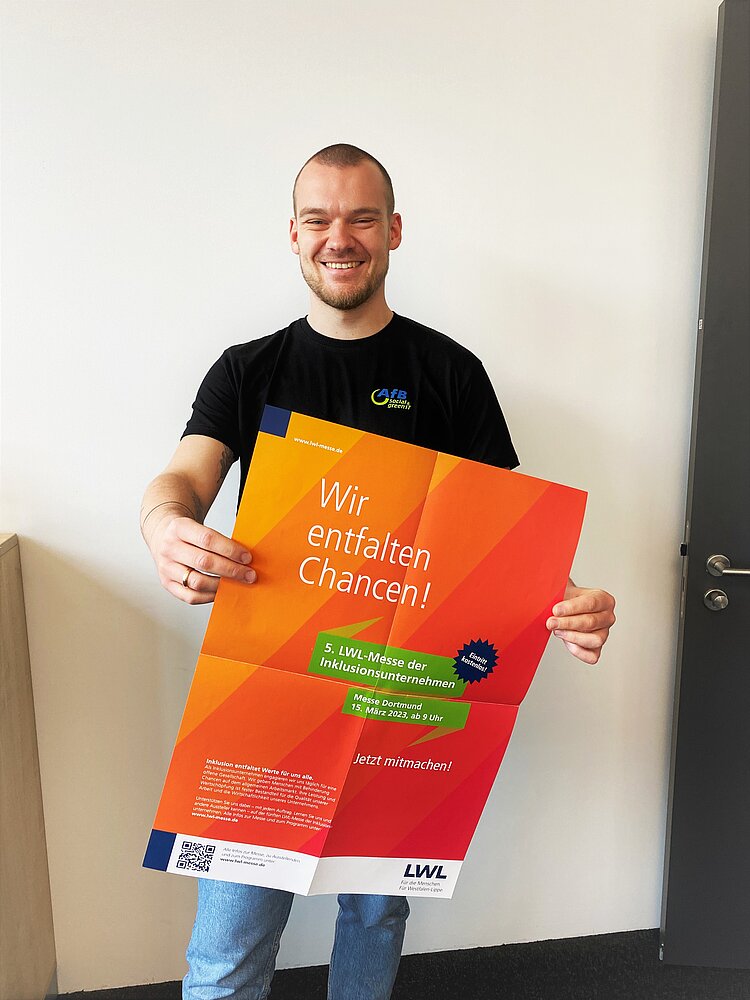 Betriebssozialarbeiter Noah Hilbrecht steht im Büro und hält das orange gefärbte Plakat der 5. LWL-Messe in Dortmund in die Kamera.