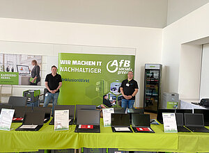 2 Männer stehen in schwarzen T-Shirts hinter einem Tisch mit grüner Tischdecke und Laptops darauf.
