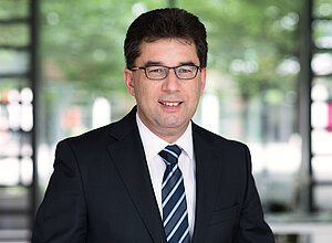 Partnermanager Thomas Richter trägt einen Anzug mit gestreifter Krawatte, eine Brille und schwarzes Haar.