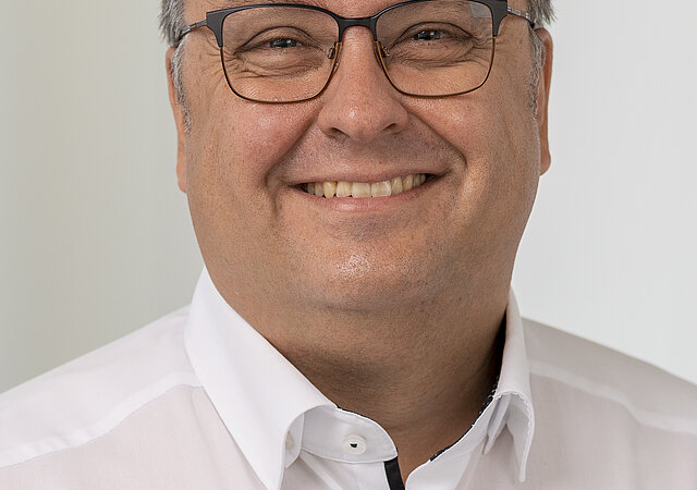 Portraitfoto von Martin Spitz. Er trägt eine Brille und ein weißes Hemd und lächelt in die Kamera.