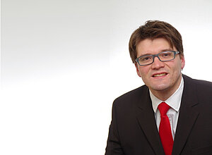 Rüdiger Imsel trägt eine Brille und einen Anzug mit roter Krawatte. Er steht vor einem weißen Hintergrund.