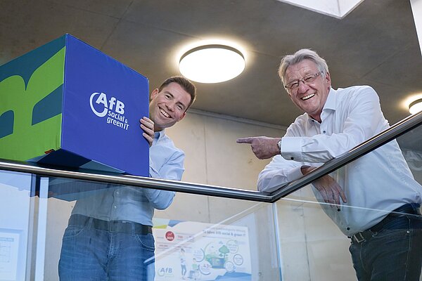 Paul Cvilak und Daniel Büchle lehnen an einem Geländer. Danie hält einen blauen Würfel mit AfB-Logo in der Hand.