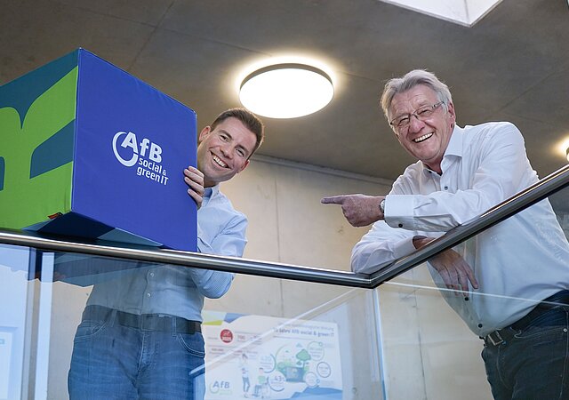 Paul Cvilak und Daniel Büchle lehnen an einem Geländer. Danie hält einen blauen Würfel mit AfB-Logo in der Hand.