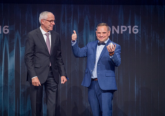 Partnermanager Tobias Negwer steht mit dem Deutschen Nachhaltigkeitspreis auf der Bühne. Er trägt einen blauen Anzug mit Fliege, freut sich und zeigt mit dem Daumen nach oben. Links neben ihm ein Mann im schwarzen Anzug.