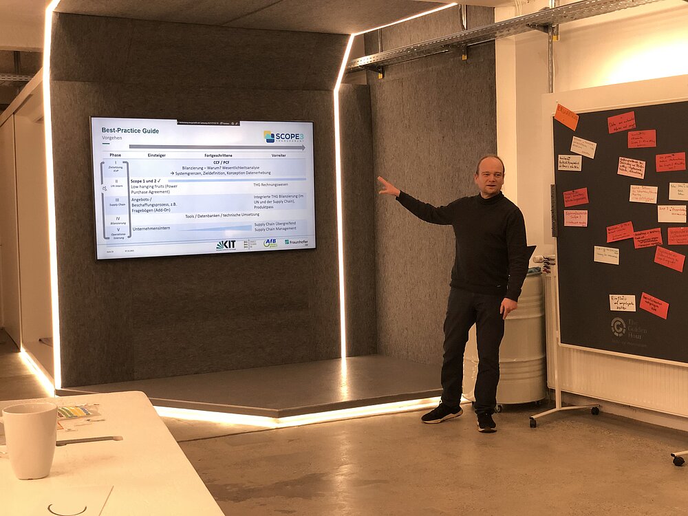 Karten Schischke informiert in einem Workshop über das Projekt Scope3transparent. Hinter ihm ist ein Monitor mit Informationen und neben ihm eine Pinnwand mit Notizen aus dem Workshop.