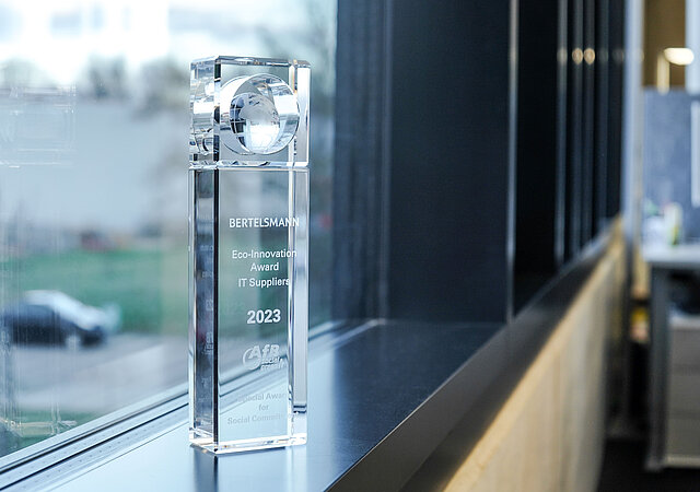 Der Bertelsmann Award steht auf einem Fensterbrett. Er ist schmal, ca. 25cm groß und aus Glas. Details zum Award sind eingraviert.