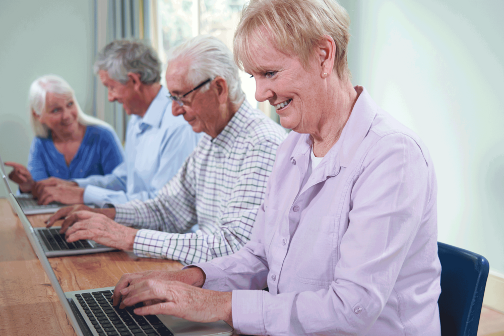 Zwei Frauen und zwei Männer über 60 Jahre sitzen an einem Tisch nebeneinander und arbeiten an zwei Laptops. 
