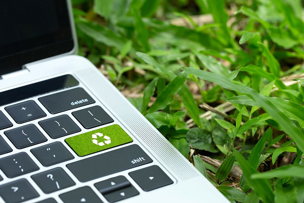 Nahaufnahme eines Laptops zwischen grünen Pflanzen. Die Enter-Taste ist grün und trägt ein Recycling-Symbol.