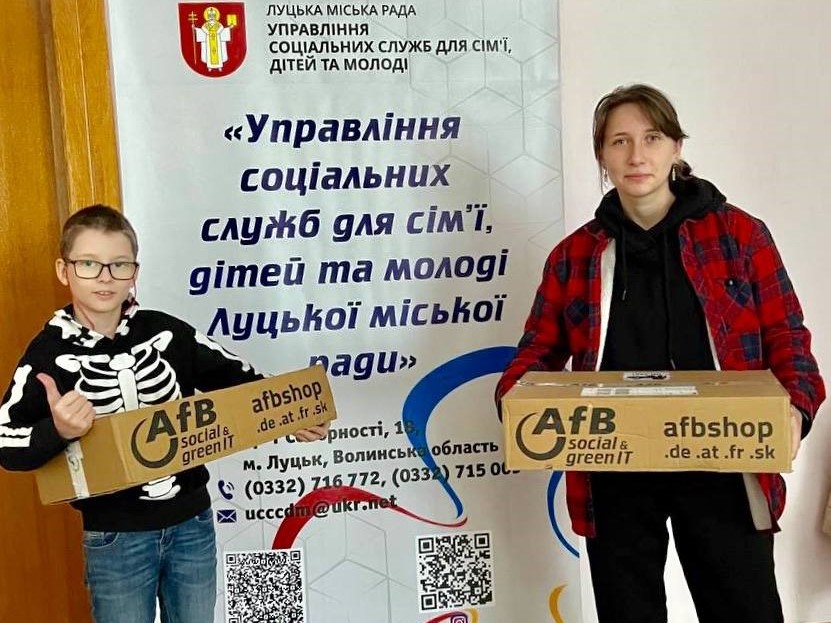Ein Junge und eine junge Frau nehmen Notebooks von AfB in Empfang. Hinter ihnen steht ein Roll-up der Verwaltung der Stadt Luzk. 
